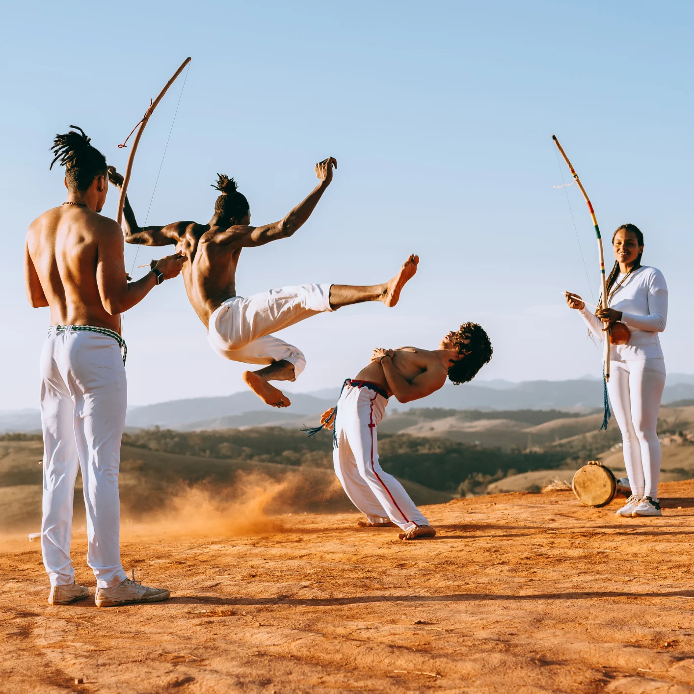 People performing capoeira outdoors, with one person mid-air while others play instruments and move in a circle.