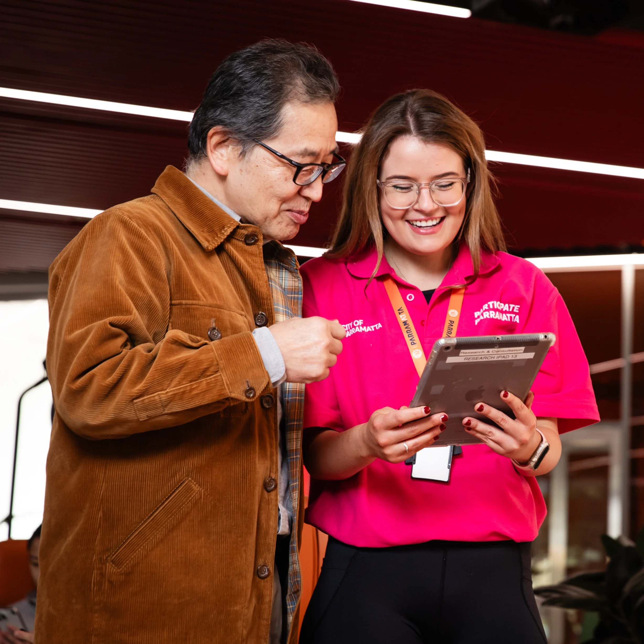 Council staff member supporting a visitor to use a tablet for digital services inside a contemporary library or community space.