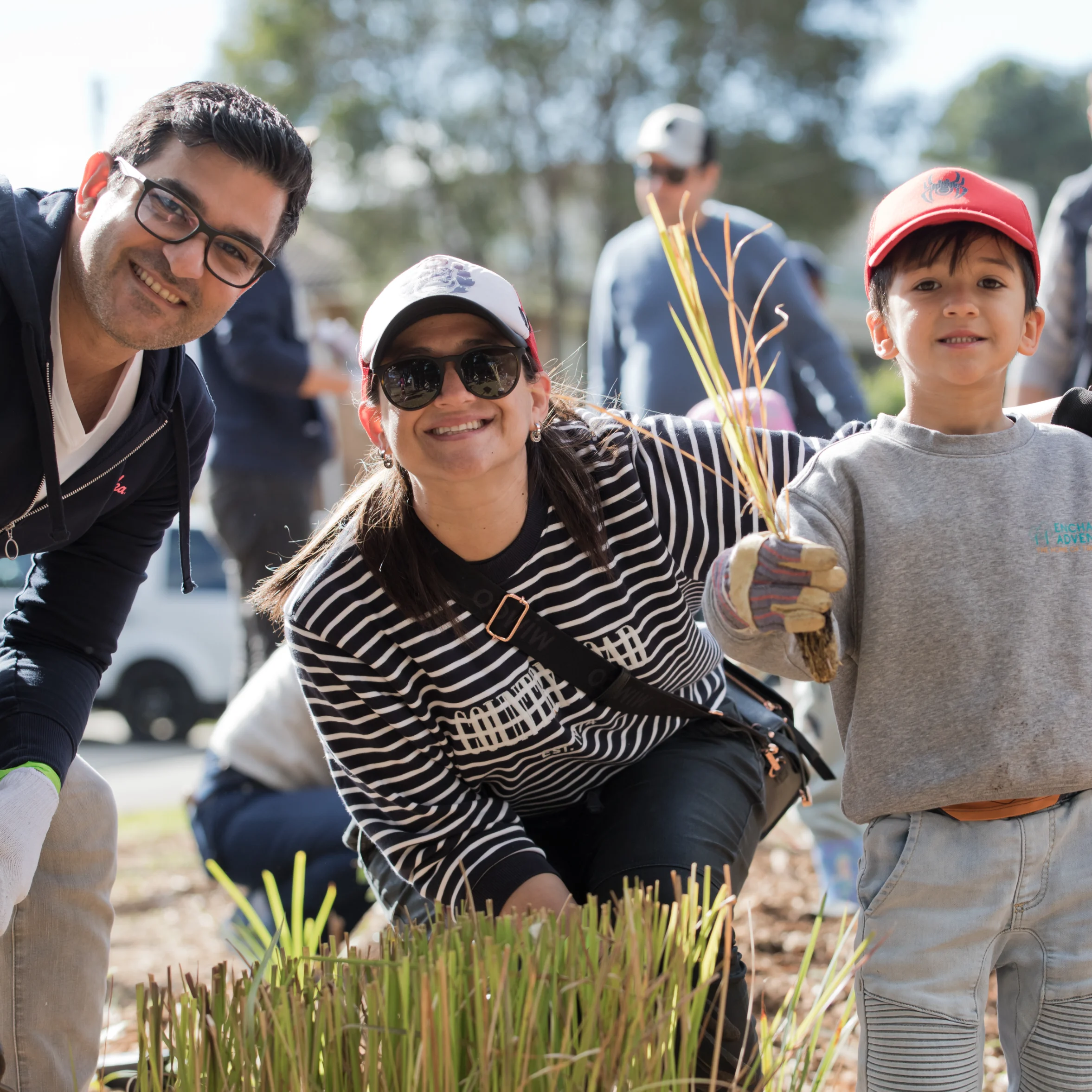 Two adults and a child smiling while planting native grasses during a community tree planting event, with others working in the background.