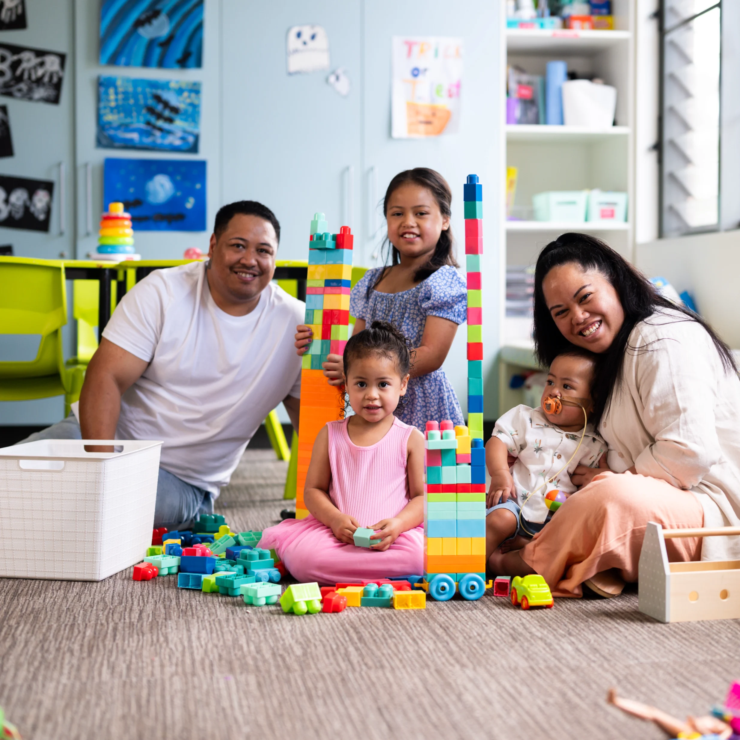 Family group with young children playing and learning together using building blocks in a childcare or community play space.