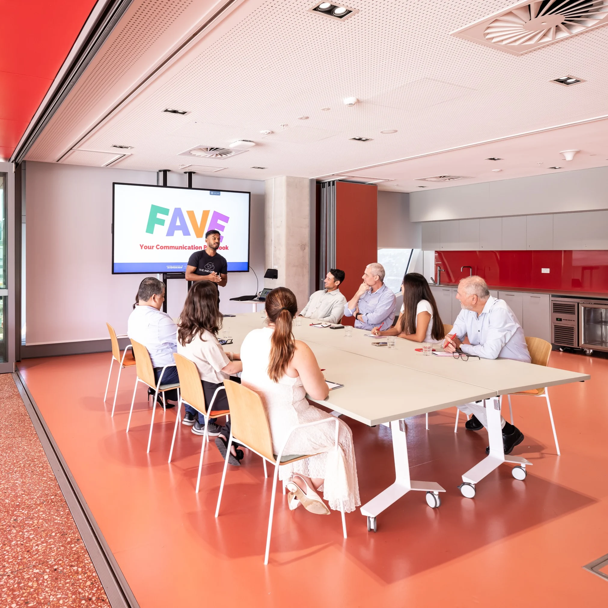 Presentation in a contemporary meeting space, with a presenter speaking to a group seated at tables and a screen showing slides.