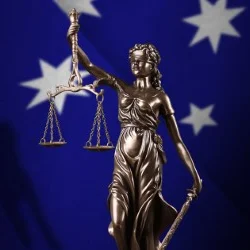 Close-up of a Lady Justice statue with scales, set against a blurred Australian flag background.
